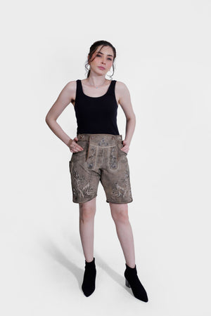 A full-body image of a woman standing in light brown Lederhosen with detailed embroidery, hands on her hips. She wears a black tank top, with a plain white background.