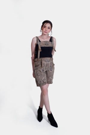 A full-body image of a woman standing in light brown Lederhosen with detailed embroidery, hands on her hips. She wears a black tank top, with a plain white background.