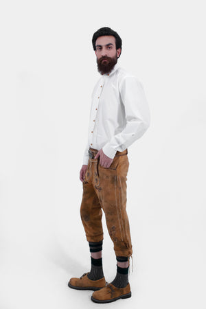 Side view of a man wearing traditional dark brown leather Bundhosen with intricate embroidery. The Bundhosen are paired with a white shirt and the outfit is accessorized with traditional leather suspenders and traditional shoes.