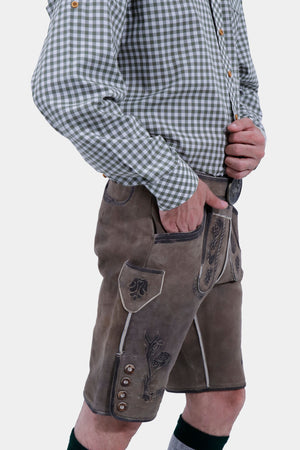 A side view of the man wearing a green checkered shirt and brown Lederhosen, highlighting the detailed embroidery. The man has his hand in his pocket.