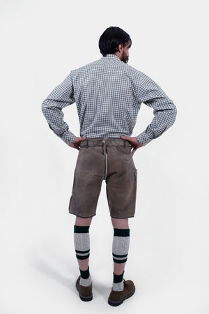 The man wearing the green checkered shirt and brown Lederhosen is shown from the back. The intricate detailing on the Lederhosen is visible, and he is wearing green knee-high socks and brown shoes.