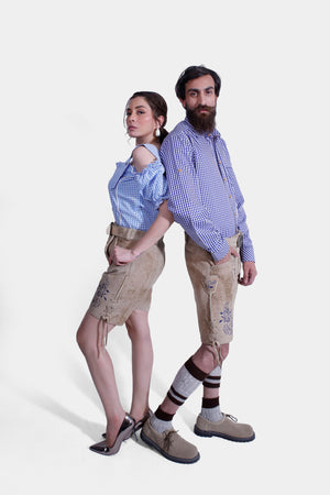 The couple posing in their Berliner Couple Outfits. The woman has one leg raised playfully while wearing a blue checkered off-shoulder blouse and embroidered light tan leather shorts. The man stands confidently beside her in a matching blue checkered shirt and light tan leather shorts with blue embroidery, completing the look with beige shoes and traditional Bavarian socks.