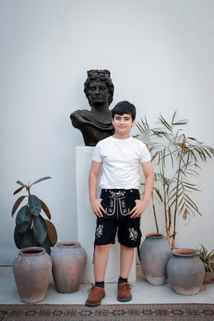 "A young boy smiling in front of a statue, wearing the 'Bernd Leno Lederhosen' in black with white embroidery, paired with a white t-shirt and brown boots.