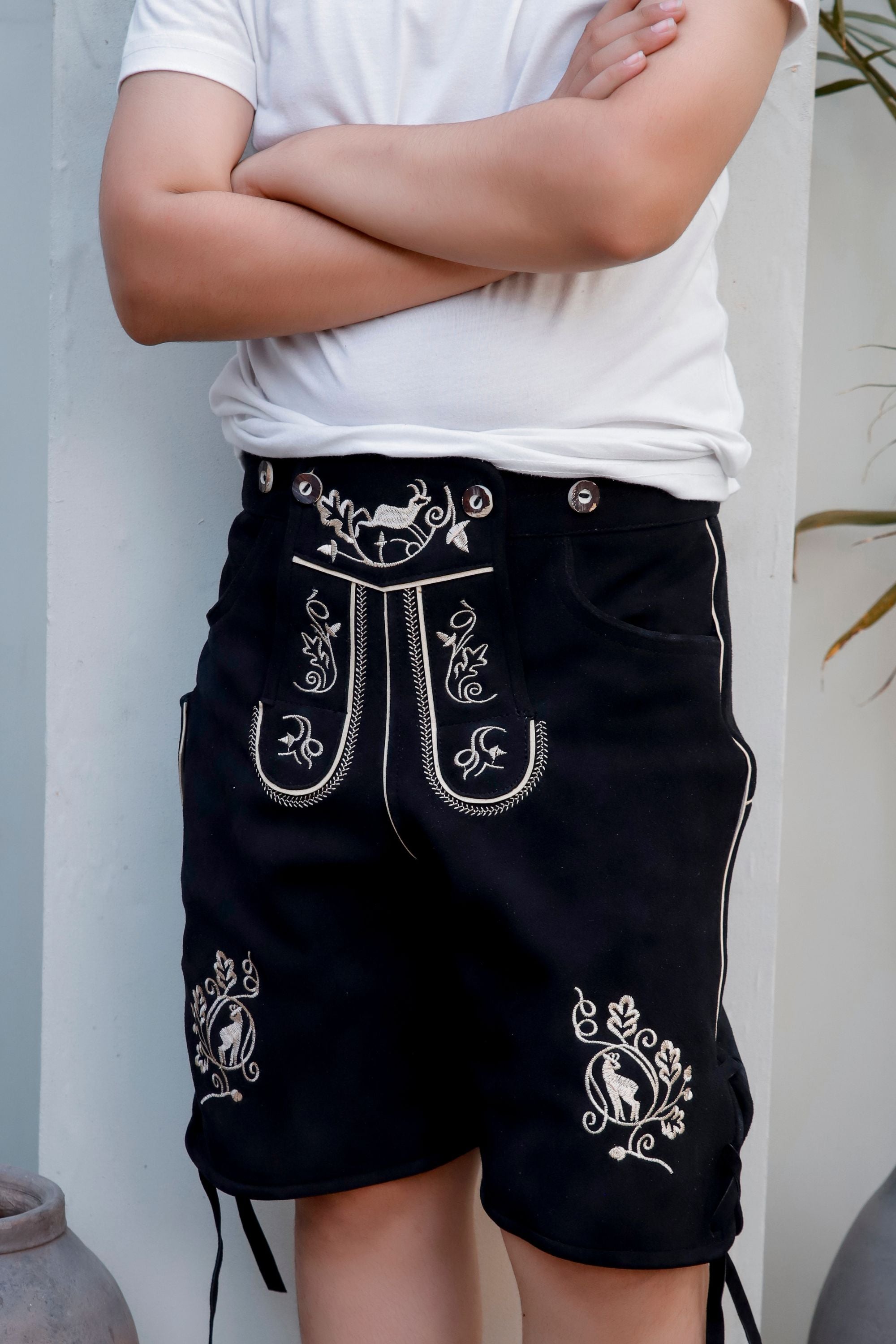 Close-up of a young boy in a white t-shirt wearing 'Bernd Leno Lederhosen' in black, showcasing detailed white embroidery on the bib and pockets, standing with arms crossed.