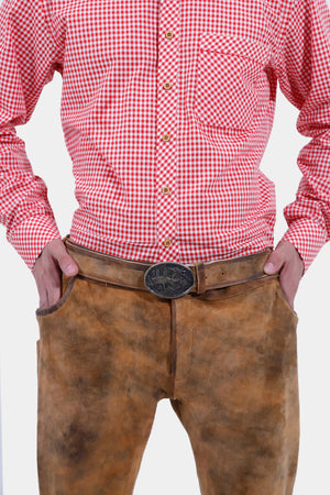 Close-up of a man wearing a red and white checkered shirt tucked into brown leather Bundhosen. The focus is on the intricate detailing of the belt buckle and the texture of the leather pants. The man's hands are in his pockets, and the shirt has wooden buttons.