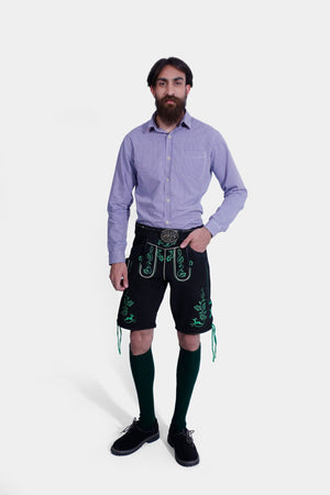 A man with a beard is standing against a white background, wearing a purple and white checkered shirt paired with black lederhosen decorated with green embroidery. He has his right hand in the pocket of the lederhosen, and is also wearing dark green knee-high socks and black shoes.