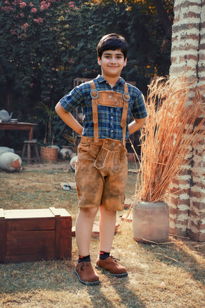 A young boy stands outdoors in a sunny garden, modeling the "Brajan Gruda Lederhosen Shorts." These traditional brown Lederhosen shorts feature natural colored embroidery on the bib and legs. He is wearing them with a plaid blue and green shirt, suspenders, and brown boots. The rustic scene includes a wooden box and tall dried grass beside a brick column, with lush greenery and flowering bushes in the background.