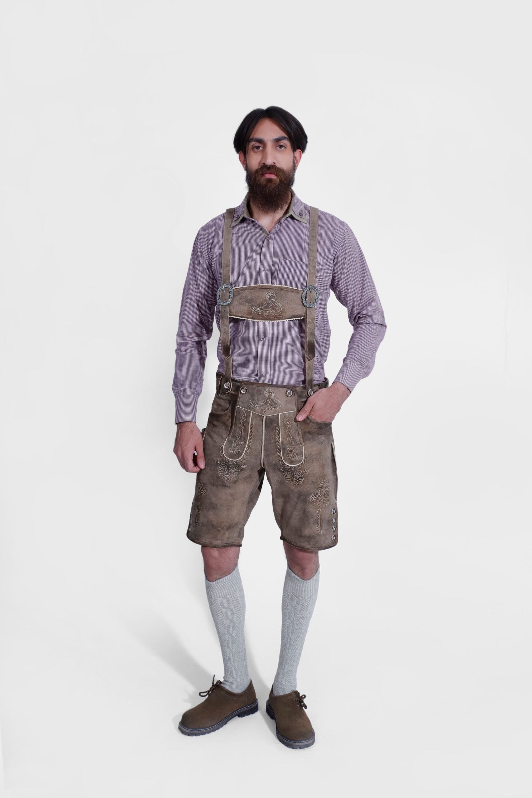 A man in "Chiemsee Castle Elite Lederhosen" with weathered brown leather and decorative suspenders, paired with a purple gingham shirt, grey socks, and brown shoes.