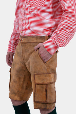 Close-up of a man wearing a red and white checkered shirt and brown leather lederhosen. The man has his right hand in the pocket of the lederhosen, showcasing the texture and details of the leather shorts.