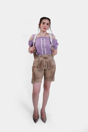 A woman modeling beige Lederhosen with detailed embroidery, paired with a purple checkered blouse and metallic high heels, showcasing a traditional Bavarian outfit.