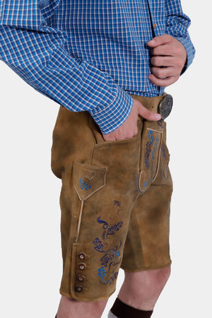 Close-up of a man wearing a blue checkered dress shirt and brown lederhosen with blue embroidery. The focus is on the detailed embroidery on the lederhosen, which includes a goat and floral patterns. The man has his left hand in his pocket and is holding the shirt with his right hand.