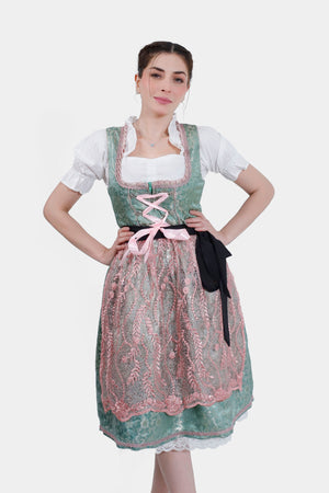 A woman wearing a green dirndl with pink lace accents, standing with her hands on her hips, looking confidently at the camera. The dirndl has a black ribbon tied at the waist and a white blouse underneath.