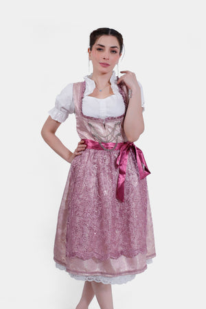 A woman models a soft pink dirndl dress with a white blouse, featuring a satin ribbon belt and lace apron, paired with metallic heels.