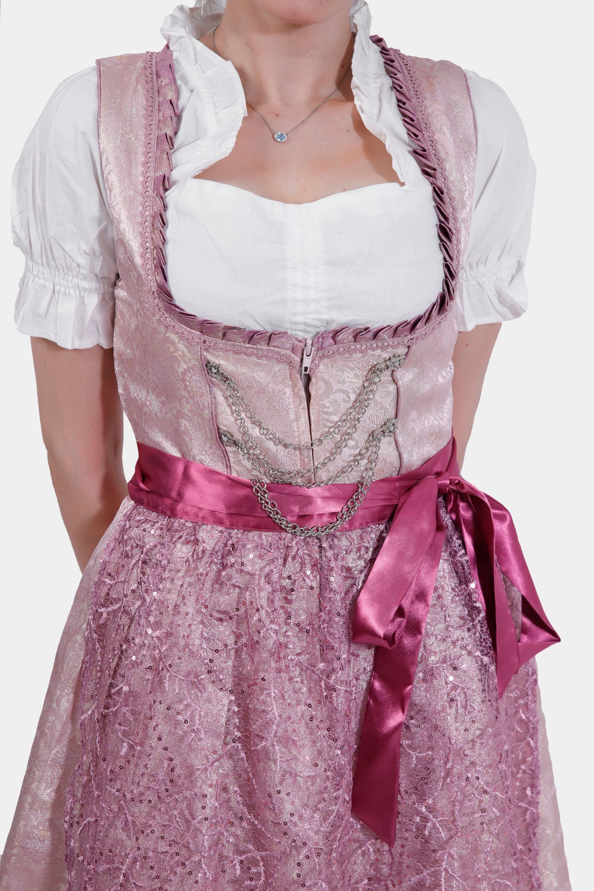 Close-up of the dirndl bodice showing intricate lace details, silver chain embellishments, and a satin pink ribbon tied around the waist.