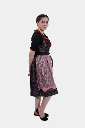A side view of the model wearing the dirndl dress, with her hand on her hip, showcasing the layered pink lace apron over the black floral skirt.