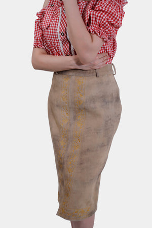 Side view close-up of the beige leather skirt with yellow embroidery. The woman's red and white checkered blouse with ruffled sleeves is partially visible as she crosses her arms.
