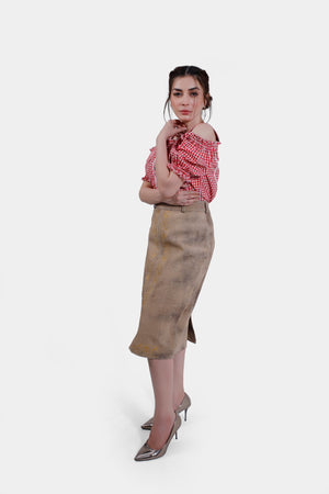 Profile view of the woman in a red and white checkered blouse and a beige leather skirt with yellow embroidery. She stands with her hand resting on her chin, looking confidently at the camera.
