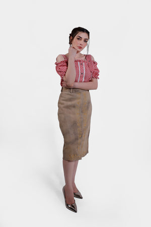 Full body shot of a woman wearing a red and white checkered blouse and a beige leather skirt with yellow embroidery. She stands with one hand under her chin, exuding a thoughtful pose.