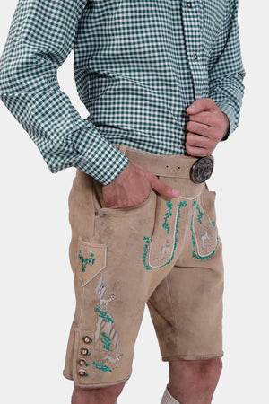 A man in tan leather lederhosen with green embroidery, his hand in his pocket, and a green and white checkered shirt, focusing on the intricate details of the lederhosen.