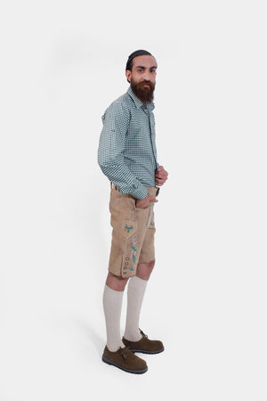 A man with a full beard wearing traditional Bavarian attire, featuring a green and white checkered shirt and tan leather lederhosen with green embroidery, standing with his hands by his sides.