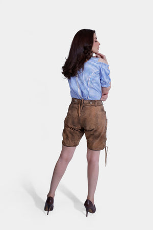 A woman with wavy brown hair wearing a blue checkered blouse and brown leather shorts, viewed from the back, standing with one arm raised.