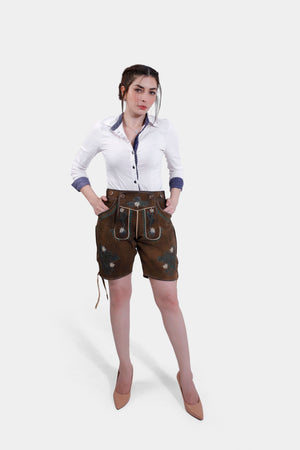 Full front view of a model in brown suede Bavarian leather shorts with green and white embroidery, standing confidently with hands in pockets, wearing a white button-up shirt and nude high heels.