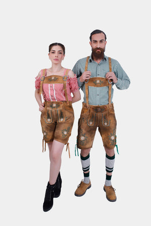 A couple wearing matching Freude Couple Lederhosen outfits. The man is in green checkered shirt and Lederhosen shorts with floral embroidery, while the woman is in a red checkered off-shoulder blouse and Lederhosen shorts with floral embroidery."