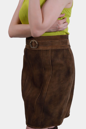 A Side view of the brown leather skirt, highlighting its detailed stitching and the ornate circular buckle on the belt. The woman's arm is partially visible, crossed over her body, emphasizing the texture and fit of the skirt.