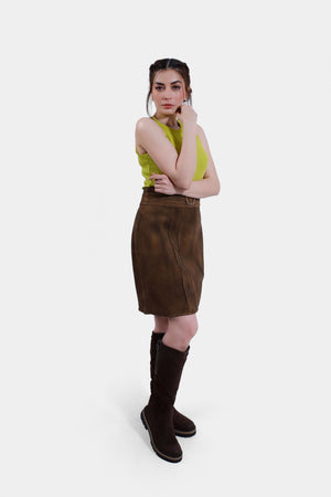 Slightly turned a body, view of the woman in the lime green top and brown leather skirt. She is also wearing knee-high brown boots, with one hand on her hip and the other resting by her side, posing confidently against a plain white background.
