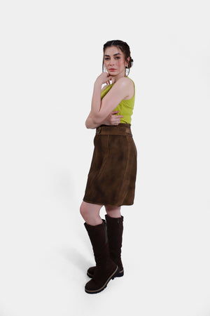 Side profile view of of the brown leather skirt, highlighting its detailed stitching and the ornate circular buckle on the belt. The woman's arm is partially visible, crossed over her body, emphasizing the texture and fit of the skirt.