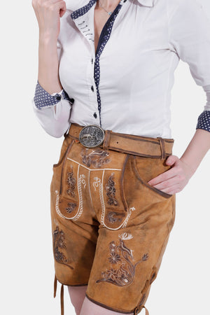 Close-up of a woman in a white blouse with blue polka dot accents and brown embroidered lederhosen. She adjusts her collar with her right hand and has her left hand in her pocket.
