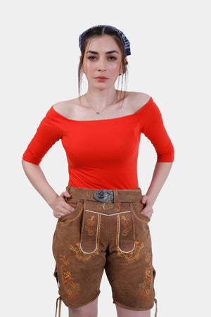 Image showing a woman wearing Heidi Klum Lederhosen in Autumn Saffron goat suede leather, paired with a bright red off-the-shoulder top. The lederhosen feature intricate embroidery and a belt with an ornate buckle.