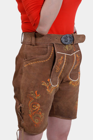 Side view of brown leather Lederhosen shorts showcasing detailed floral embroidery, a brown belt with a silver buckle, and lacing on the side, paired with a bright red top.