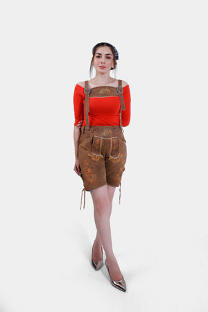 Full-body view of a model wearing traditional brown leather Lederhosen shorts with orange floral embroidery, suspenders, and a red off-the-shoulder top, paired with silver high heels.