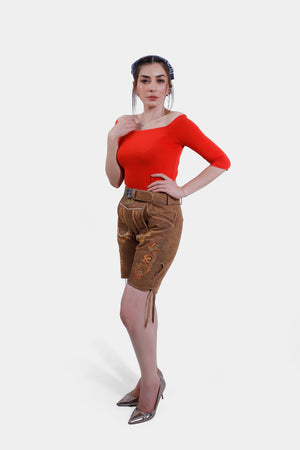 Full-body view of a model in brown leather Lederhosen shorts with orange embroidery, a matching belt, and a red off-the-shoulder top, accessorized with a headscarf and silver high heels.