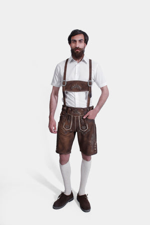 A man with a full beard stands against a white background, dressed in "Hudson Valley Lederhosen." These knee-length Lederhosen are made of weathered brown leather and feature ornate silver embroidery. He wears them with a crisp white shirt, grey knee-high socks, and dark brown shoes, showcasing a traditional yet sophisticated Bavarian style.