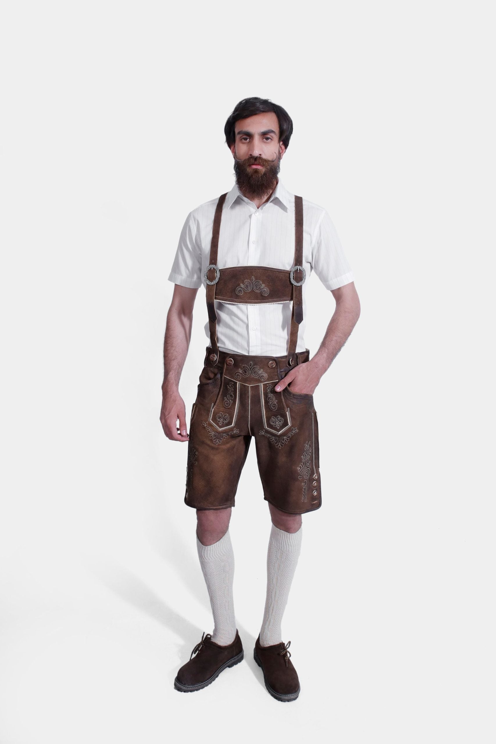 A man with a full beard stands against a white background, dressed in "Hudson Valley Lederhosen." These knee-length Lederhosen are made of weathered brown leather and feature ornate silver embroidery. He wears them with a crisp white shirt, grey knee-high socks, and dark brown shoes, showcasing a traditional yet sophisticated Bavarian style.