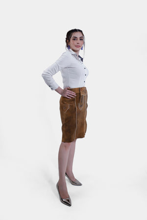 Side profile of a woman wearing a white shirt and a brown leather Jennifer Lederhosen Skirt with intricate embroidery, standing with one arm crossed and her hand near her chin.