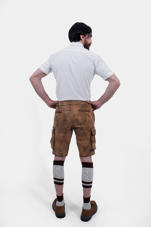 A man with a beard is standing with his back to the camera, hands on hips, wearing a short-sleeved white shirt and brown leather lederhosen. He is also wearing brown and grey knee-high socks and brown shoes. The lederhosen has pockets on both sides and is detailed with stitching.