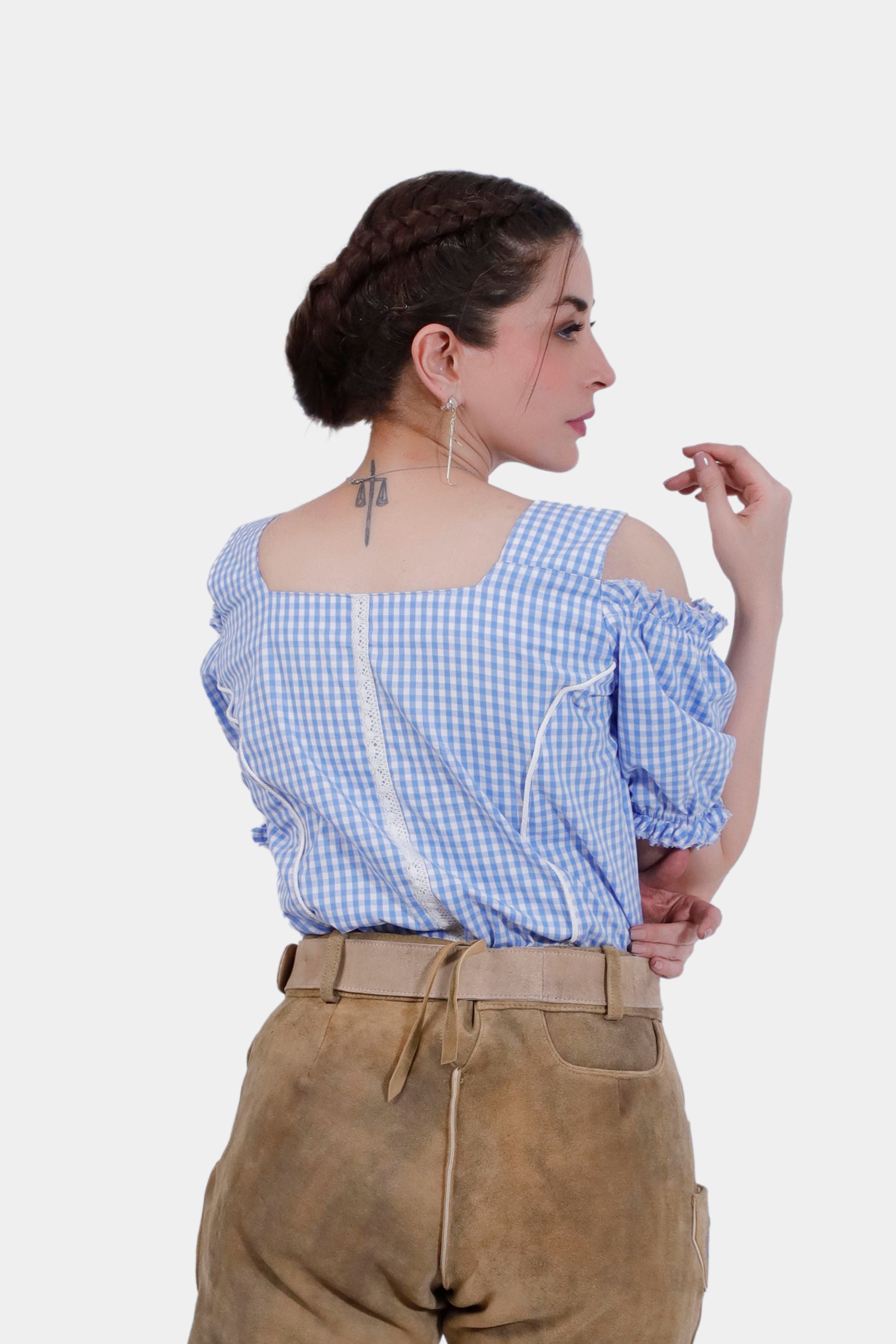 A woman is shown from the back wearing a blue checkered off-shoulder traditional blouse with ruffled sleeves and a lace trim down the back. She pairs the blouse with tan leather lederhosen. Her hair is styled in a braided updo, and she has a tattoo of scales on the back of her neck.