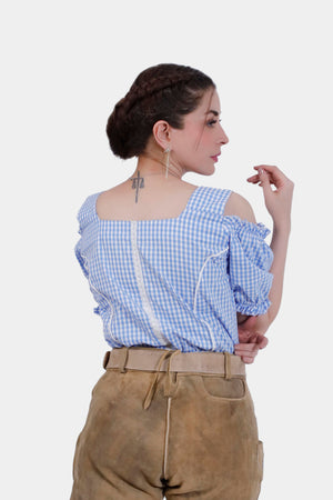 A woman is shown from the back wearing a blue checkered off-shoulder traditional blouse with ruffled sleeves and a lace trim down the back. She pairs the blouse with tan leather lederhosen. Her hair is styled in a braided updo, and she has a tattoo of scales on the back of her neck.