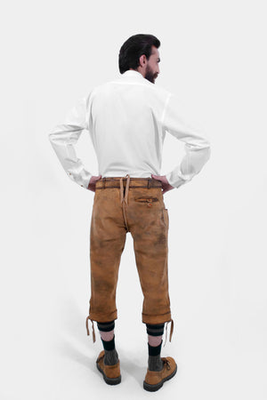 Rear view of a man wearing traditional brown leather Bundhosen and a white shirt. The image showcases the back details of the Bundhosen, including the tie straps and embroidery on the back pocket. The man is also wearing traditional Bavarian socks and shoes.