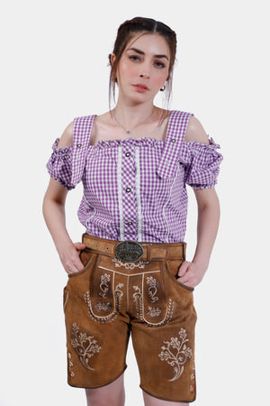Model wearing traditional brown leather lederhosen shorts paired with a purple checkered blouse with off-the-shoulder sleeves and a decorative belt.