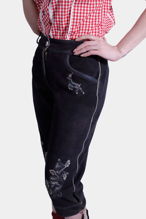 A detailed view of the woman's black lederhosen pants, emphasizing the intricate embroidery of a deer and floral patterns on the fabric. Her red and white checkered blouse is partially visible, with her hand on her hip.
