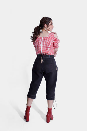 A back view of the woman wearing a red and white checkered off-shoulder top with a back zipper, paired with black lederhosen pants. The pants feature subtle embroidery and are cropped just below the knees. She completes the look with red ankle boots, and her hair is styled in a loose ponytail.