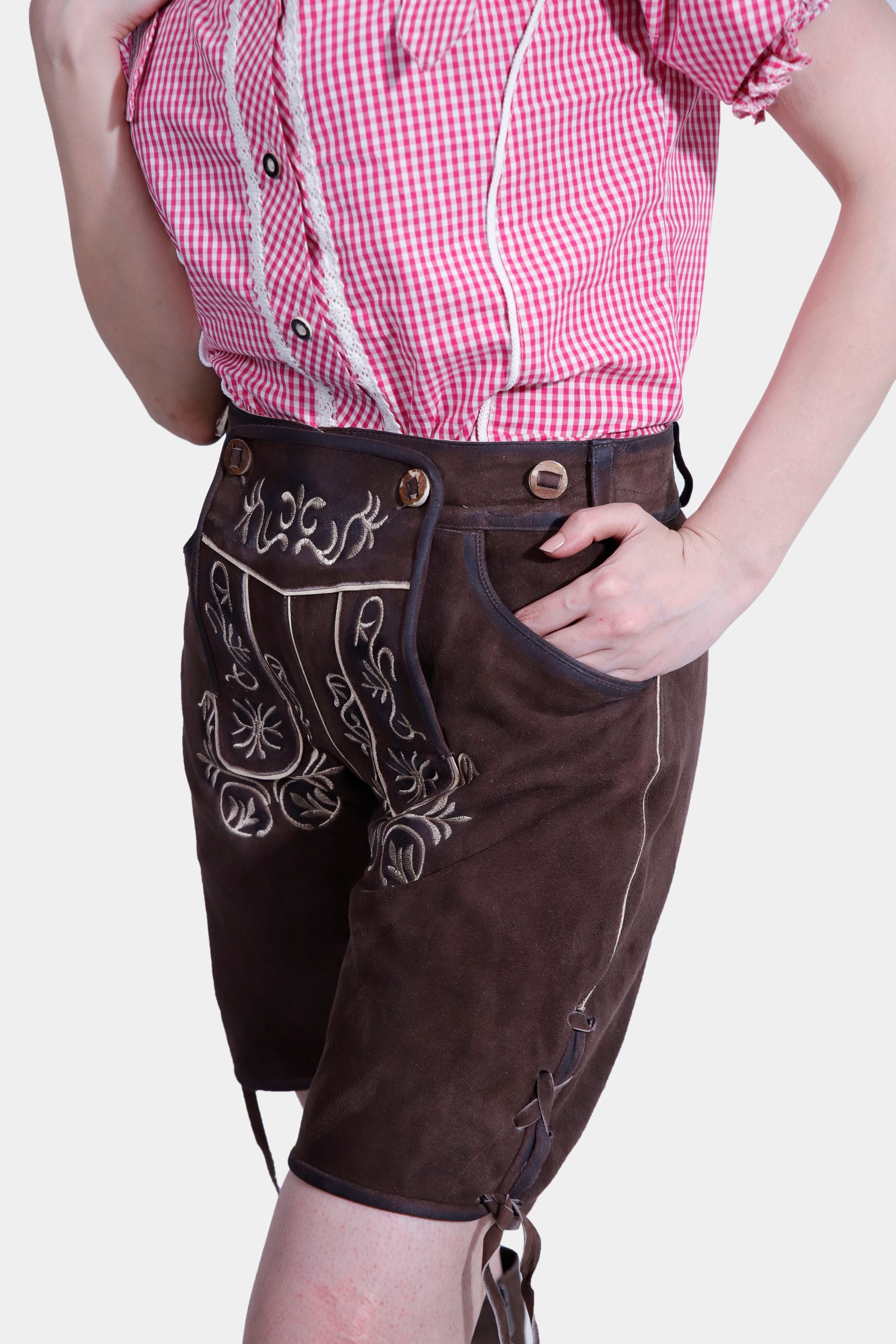A close-up view of the woman's outfit, focusing on the brown lederhosen shorts with detailed floral embroidery and buttons. She is wearing a pink and white checkered off-shoulder blouse, and her hand rests inside the pocket of the shorts.