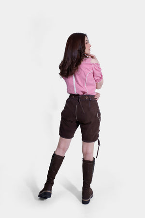 The woman is standing with her body slightly turned, showing the back of her outfit. The brown lederhosen shorts and the back of the pink and white checkered blouse with a zipper detail are visible. She is wearing knee-high brown boots, and her hair is styled in loose waves.
