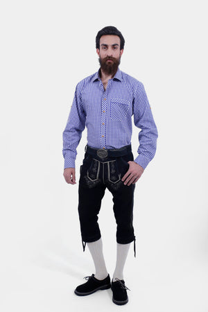 A man with a beard and styled hair wearing a blue and white checkered shirt paired with black leather bundhosen and cream-colored knee-high socks, standing with his hands by his sides and looking directly at the camera.