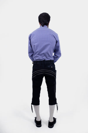 Rear view of a man wearing a blue and white checkered shirt and black leather bundhosen, showing the back pockets and overall fit of the bundhosen along with cream-colored knee-high socks.