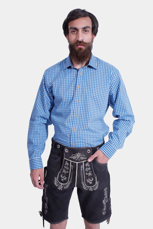A man in Mountain Majesty Lederhosen with a blue checkered shirt, standing with hands in pockets and a neutral expression.